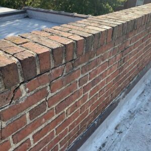 A detailed close-up photograph of a weathered brick parapet wall on a rooftop, showing crumbling mortar and cracks under natural light.