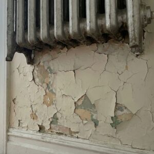 Peeling and cracked paint layers on an interior wall next to an old radiator in an older apartment building.